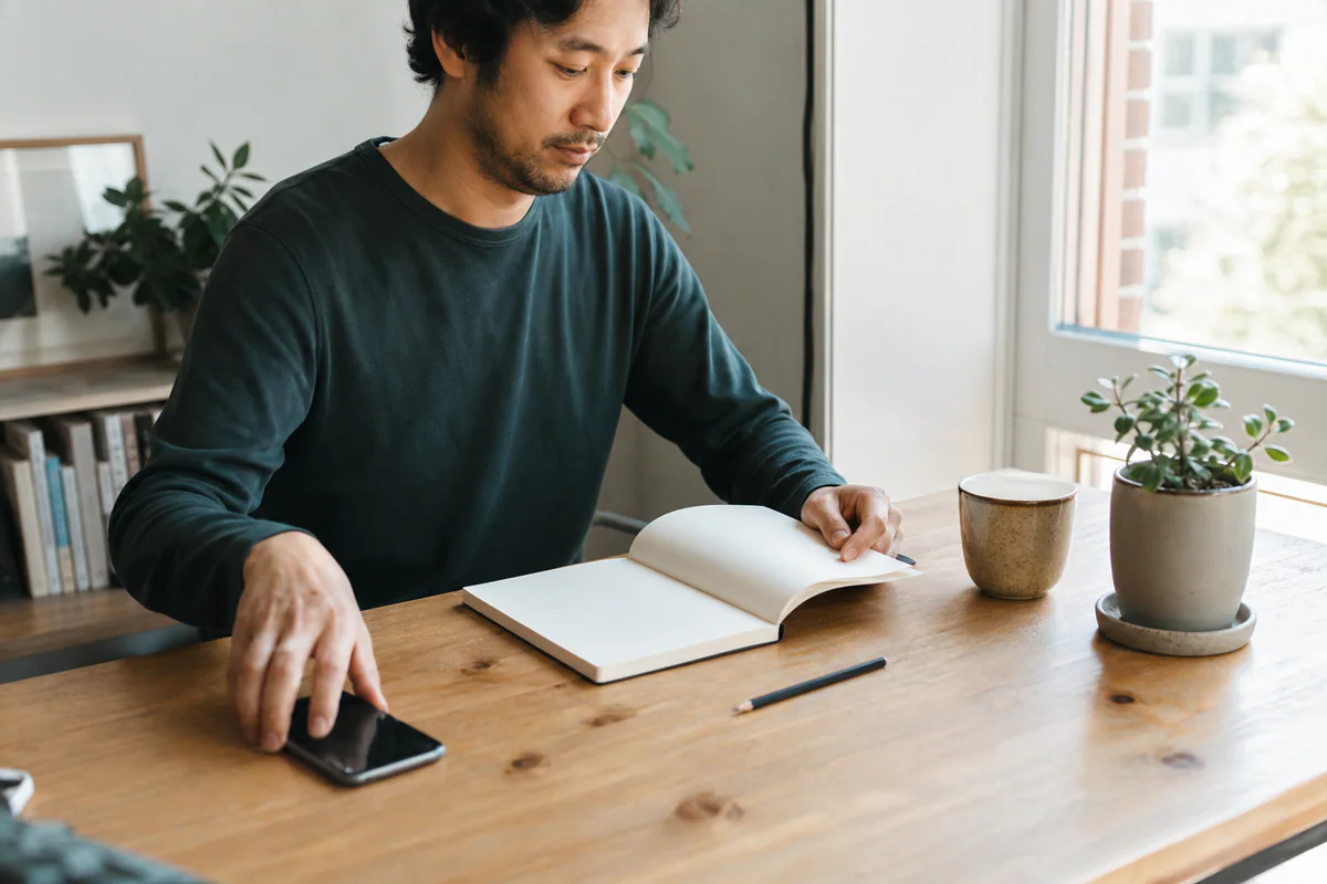 A person at a sunlit desk placing a phone face down and opening a notebook to one simple task.