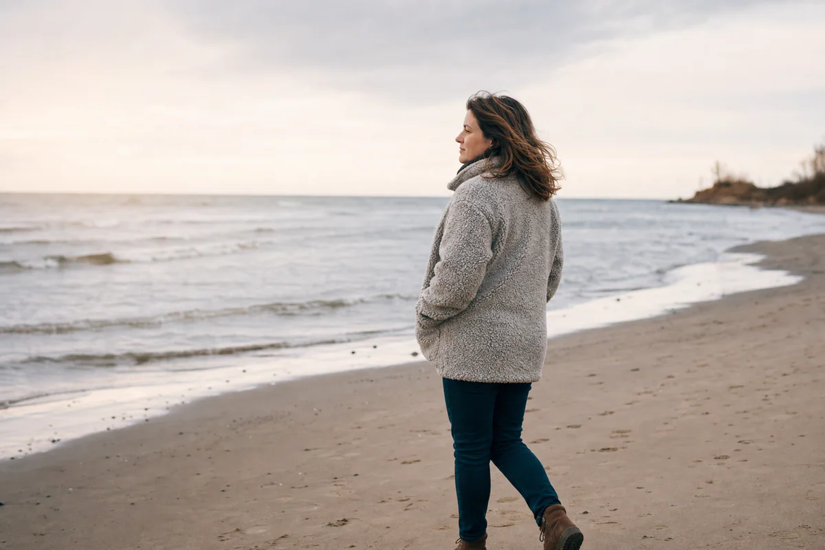 A person walking slowly along a calm shoreline, wrapped in a textured jacket and looking toward the water.