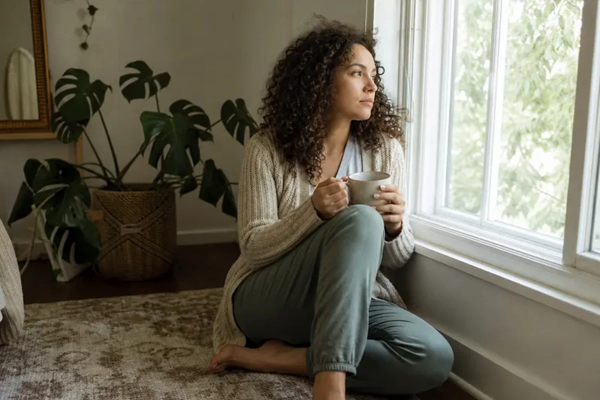 A person standing by a bright window, resting one hand lightly near the chest and noticing a slow breath.