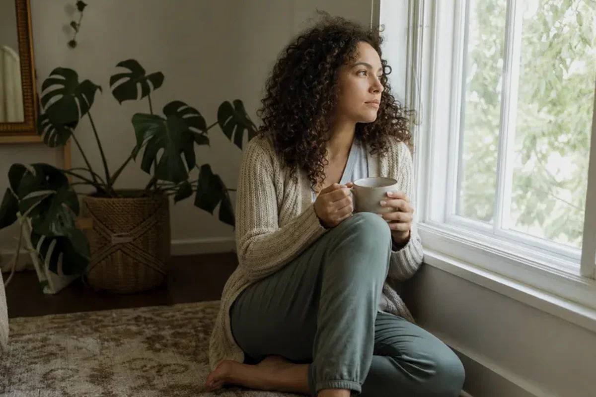 A person standing by a bright window, resting one hand lightly near the chest and noticing a slow breath.