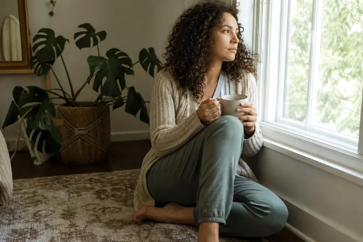 A person standing by a bright window, resting one hand lightly near the chest and noticing a slow breath.