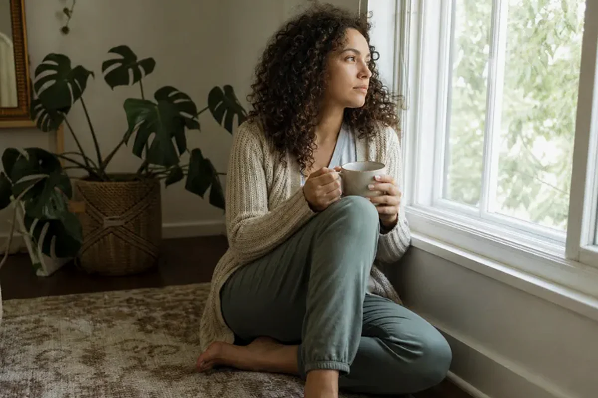 A person standing by a bright window, resting one hand lightly near the chest and noticing a slow breath.